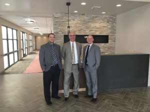 Image of three smiling men in suits and ties standing in front of a reception counter at a Cascadia facility.