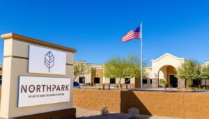 Main entrance of NorthPark Health and Rehabilitation. Two-tone tan stucco exterior with brick. An American flag waves in the breeze at the edge of the entrance. A brown brick fence sits just behind a large sign displaying the NorthPark logo.