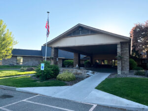 Exterior view of the main entrance to Twin Falls Transitional Care. A brown stone building with a sidewalk leading to a drive-through portico. Grass, deciduous trees, shrubs and gravel beds surround the sidewalk with an American flag flying from a tall pole. Twin Falls Transitional Care Of Cascadia